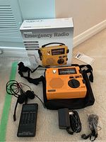 Photo showing the Grundig FR200G emergency radio, its box with product image and information, a black carrying strap, power adapter, and an accompanying small black Realistic Stereo Mate device with earbuds attached.