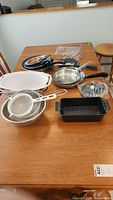 Overview of kitchen items on wooden table showing 7 frying pans stacked, assorted strainers including metal and plastic, a white ceramic baking dish, black perforated meatloaf pan and metal baking racks.