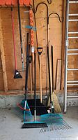 Full view of garden tools including rakes, shovels, brooms, and shepherd's hooks hung and leaned against the wall.
