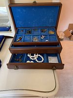 Jewelry box open showing blue velvet lined compartments with assorted jewelry items, including cufflinks, pins, and chains in the top tray and various bracelets and earrings in the lower drawer.