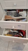 Two open drawers showing a variety of kitchen utensils including spatulas, measuring cups, and knives in the top drawer and graters, peelers and juicer in the bottom drawer.