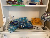 Shelf display containing assorted glass platters and bowls, decorative magnets, and a Tiffany-style candle lamp.