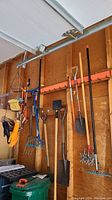 Multiple garden tools hanging on hooks on a wooden wall including a hoe, several rakes, shovels, a manual edger with star-shaped spikes, and other hand tools.
