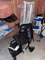 Photo showing the fold-up walker with padded black seat and backrest, storage basket below, and adjustable crutches wrapped in plastic behind it.