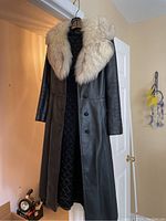Front view of full length black leather coat hanging on a door, showing large cream fur collar and quilted black lining interior