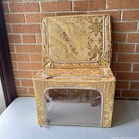 Two stackable collapsible storage boxes, one upright and one stacked on top, showing gold damask pattern with clear plastic window zipper compartment.