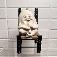 Front view of the anthropomorphic egg figurine seated on a black wooden chair with woven seat against white brick background.