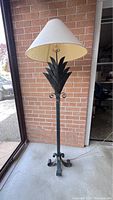 Full view of the floor lamp standing upright near a window and brick wall background, showing the beige conical lampshade and palm frond design on the metal stand.