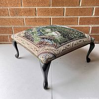 Side angle view of the foot stool showing the tapestry upholstered padded top and one of the black wood cabriole legs.
