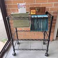 Front-left angle view of black metal rolling filing cart with blue hanging file folders, wooden Porta-File lock box, green metal supply box, and box of ColGlo ruled index cards on top shelf