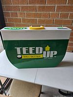 Closed view of the Teed Up Folding Golf Ball Setter showing green and white casing with yellow handle and logo.