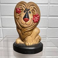 Front view of the ceramic lion pencil holder showing detailed painted features including red flowers by eyes, black nose, tan mane and paws, on black base.