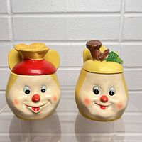 Two vintage anthropomorphic ceramic wall pockets shown from front, smiling faces with lids featuring flower and log decorations.