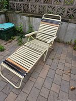 Photo of vintage cream-colored metal frame chaise lounge with slatted seat and back, high back design, wooden slats at headrest and footrest, showing rust and wear, placed on outdoor stone patio near concrete wall