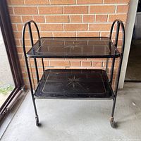 Front view of vintage black metal fold away serving tray cart showing two starburst trays and tubular handles.