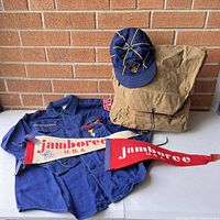 Photo of Boy Scout backpack, Cub Scout shirt, navy blue cap, and two Jamboree pennants displayed on a table with brick background.