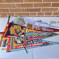 Photo showing a collection of colorful vintage souvenir pennants arranged on a cardboard surface against a brick wall. Visible designs include Acadia, 1000 Islands Museum, Cape May Ferry, Gettysburg, and St. Lawrence Seaway.