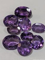 Nine faceted amethyst gemstones in oval and round shapes, showing their purple color and clarity on a white surface.