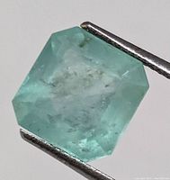 Close-up image of a single loose emerald being held by metal tweezers showing its octagonal step-cut and internal inclusions.