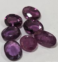 Seven faceted oval-shaped ruby gemstones displaying their color and cut under lighting, highlighting internal inclusions and clarity.