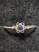 Close-up photo of a silver ring with a blue round stone set in a flower-shaped bezel on a grey background.