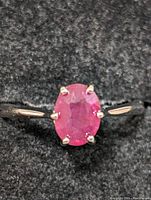 Close-up photo of sterling silver ring with oval ruby gemstone on black fabric background, showing the front and top view of the ring, highlighting the ruby and prong setting.