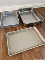 Photo showing three aluminum baking pans on a wooden floor: a ribbed cookie sheet (10x15), a small pan (9x12), and a large pan (10x15) with handles.