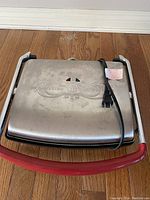 Image of Bella Cucina sandwich press with stainless steel lid and red handle, power cord attached