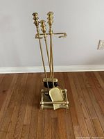 Photo of brass fireplace tool set with poker, brush, shovel hanging on brass stand on wooden floor against wall.
