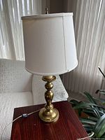 Full view of the brass table lamp on a wooden side table next to a couch and plant, showing the lamp with its plain white tapered fabric shade under natural light from a window.