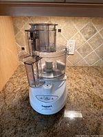 Front view of Cuisinart Prep 9 food processor on granite countertop with tiled backsplash. Unit is white with clear plastic work bowl and feeding tubes, showing buttons and blades inside.