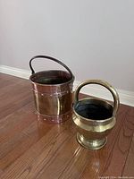 Photo showing two brass buckets on wooden floor against light wall. Larger bucket to left with riveted bands and handle, smaller bucket to right with rounded base and handle.