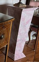 Photo shows a tall wooden pedestal painted with a pink marble-like finish, tapered square shape with stepped base, placed indoors beside furniture.
