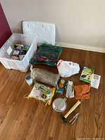 Photo of assorted survival and emergency kit items arranged on hardwood floor including hatchet, bedroll, mess kit, gloves, flashlight, storage box and various supplies.