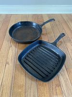 Photo shows two cast iron pans on wooden floor: a round fry pan and a square grill pan with handle holes, both with signs of use.
