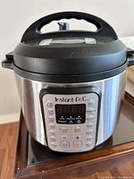 Front view of Instant Pot showing stainless steel body, black lid with handle, and digital control panel with multiple cooking options.