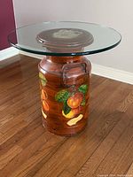 Milk can table with orange fruit painting and glass tabletop shown in room with wood floor and mauve wall.