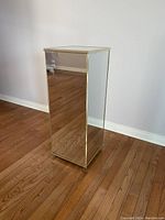 Front view of a rectangular mirrored display column with gold trim standing on a wooden floor against a gray wall.