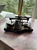 Photo of an old postal scale with balance beam, two platforms for weighing, and several cast iron weights on a wooden base near a window.