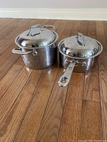 Photo showing two stainless steel sauce pans with lids on hardwood floor. Larger pan has two side handles, smaller pan has one long handle.