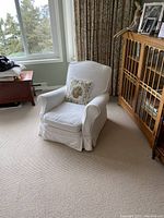 White upholstered armchair covered with white slipcover, positioned in a room beside a large window and wood cabinet. Needlepoint cushion on chair is visible but excluded from lot.