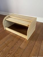 Wooden rolling top breadbox on hardwood floor next to white wall, showing curved rolling slat lid and open front.