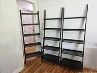 Three black ladder shelves arranged side by side with five shelves each, shown leaning against a white wall on a wooden floor.