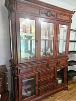 Front view of large wooden china cabinet with three glass doors on upper section and drawers and cabinets below. Ornate carved wood details and medium brown finish visible.