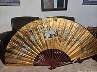 Front view of the large oriental decorative fan resting on couch, showing full expanded fan with gold paper featuring crane and blossom design.