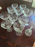 Eleven pieces of assorted glassware on dark wood surface, mainly stemmed coupe glasses with various etched and cut designs plus a glass bottle with stopper.