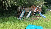 Photo showing entire wooden playground set with three slides, two swings under an A-frame, seesaw platform, rock climbing wall, multiple ladders, and a hanging red punching bag. Located outdoors in a grassy shaded yard.