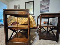 Two wooden end tables with dark wood finish, rectangular tops, middle and bottom shelves, cross-braced legs, placed on carpeted floor.