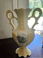 Front view of ceramic vase showing one scenic cottage painting, lattice texture, dual ornate handles, and flared scalloped base.