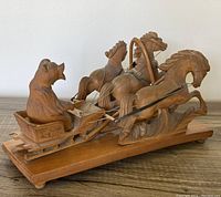 Side angled view of the wooden Troika sculpture showing the bear figure driving three horses on a sleigh mounted on a wooden base.